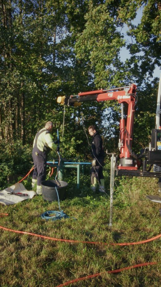 Zwei Arbeiter bedienen einen robotergesteuerten Kran, umgeben von Bäumen und Gras, und nutzen im Freien Schläuche und Werkzeuge.