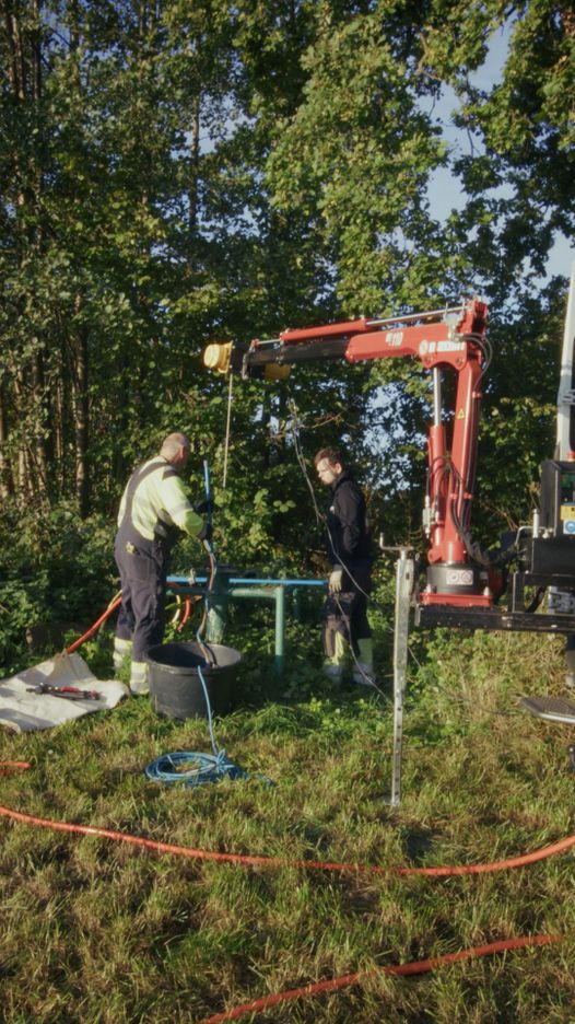 Zwei Arbeiter bedienen einen robotergesteuerten Kran, umgeben von Bäumen und Gras, und nutzen im Freien Schläuche und Werkzeuge.