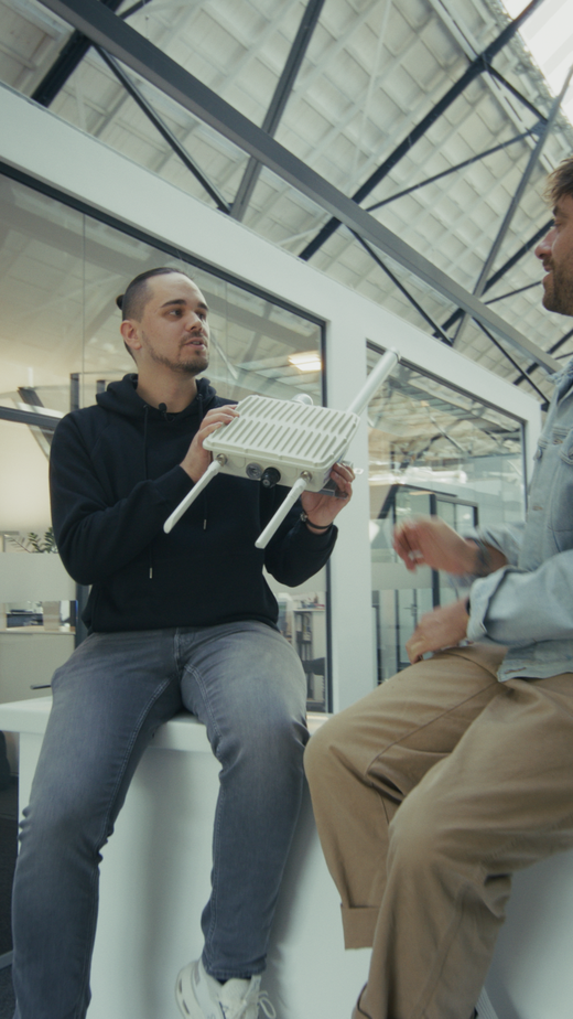 Zwei Menschen im Gespräch; einer sitzt in einem modernen Büro und hält ein weißes elektronisches Gerät mit Antennen in der Hand.