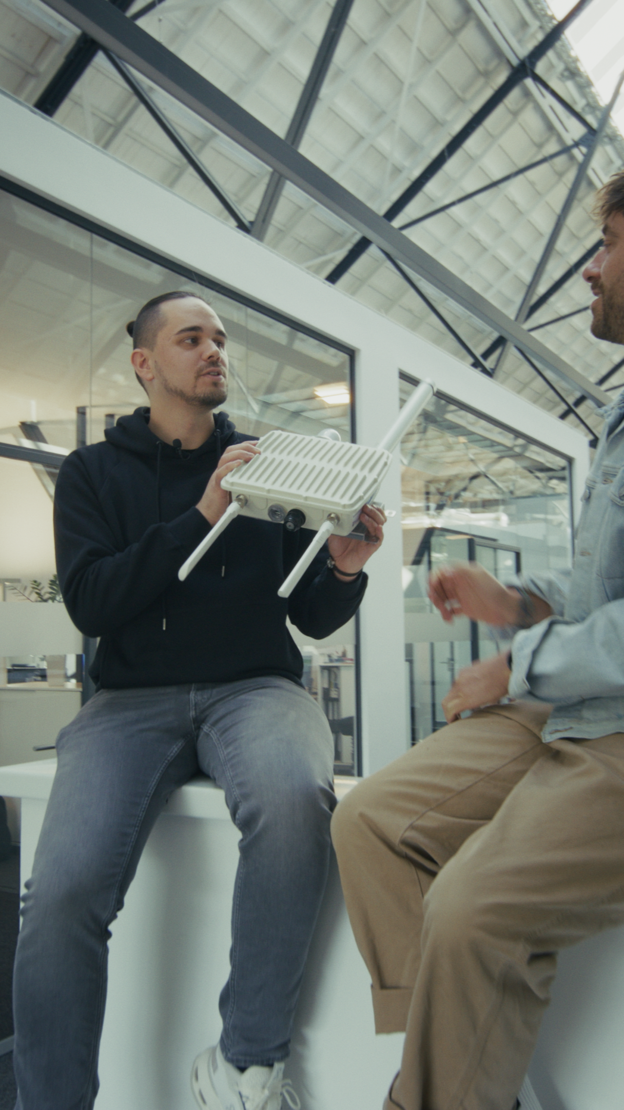 Zwei Menschen im Gespräch; einer sitzt in einem modernen Büro und hält ein weißes elektronisches Gerät mit Antennen in der Hand.