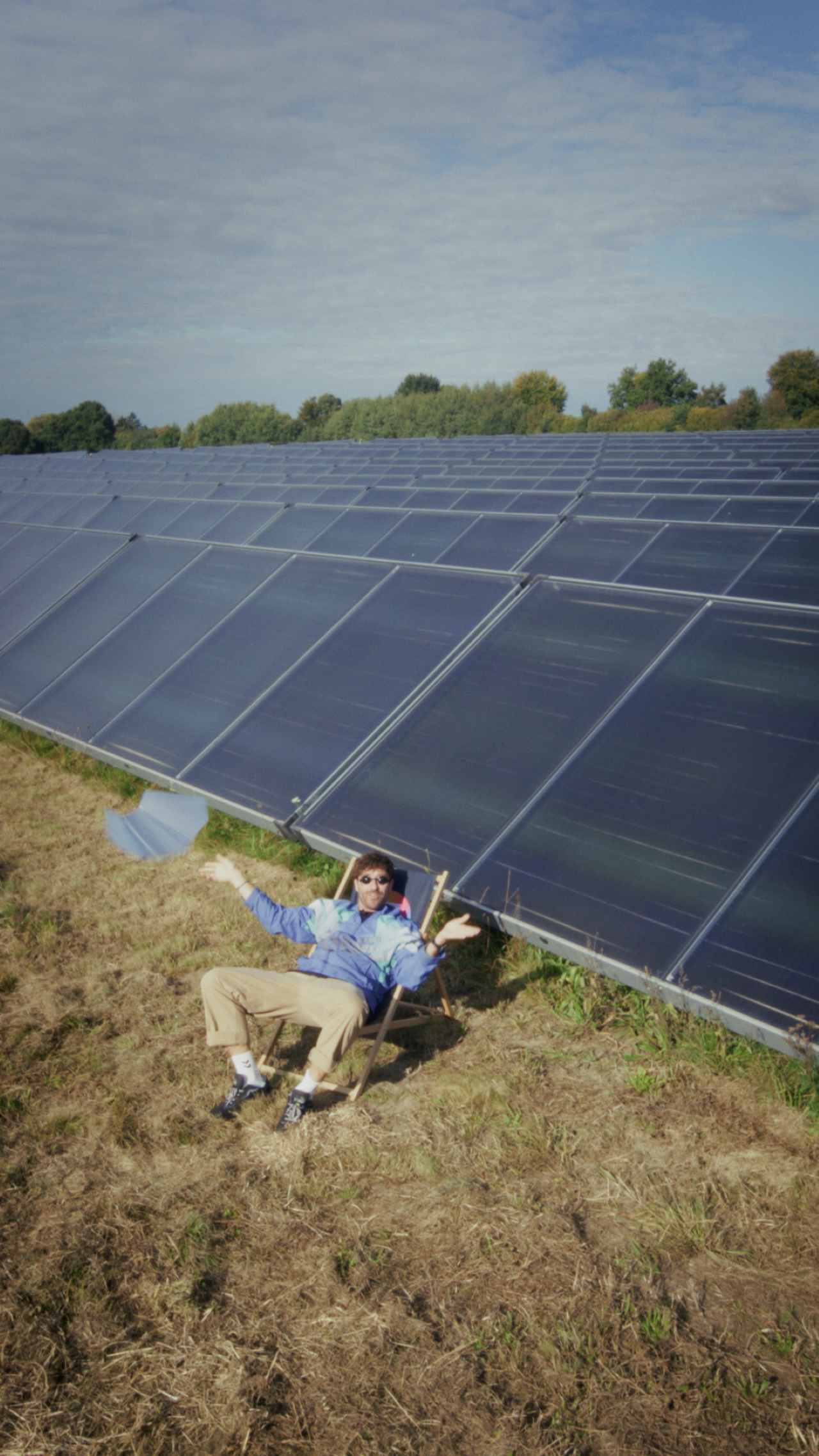 Eine Person, die an einem sonnigen Tag in einem Sessel neben einer großen Solaranlage sitzt, mit teilweise bewölktem Himmel und Bäumen im Hintergrund.