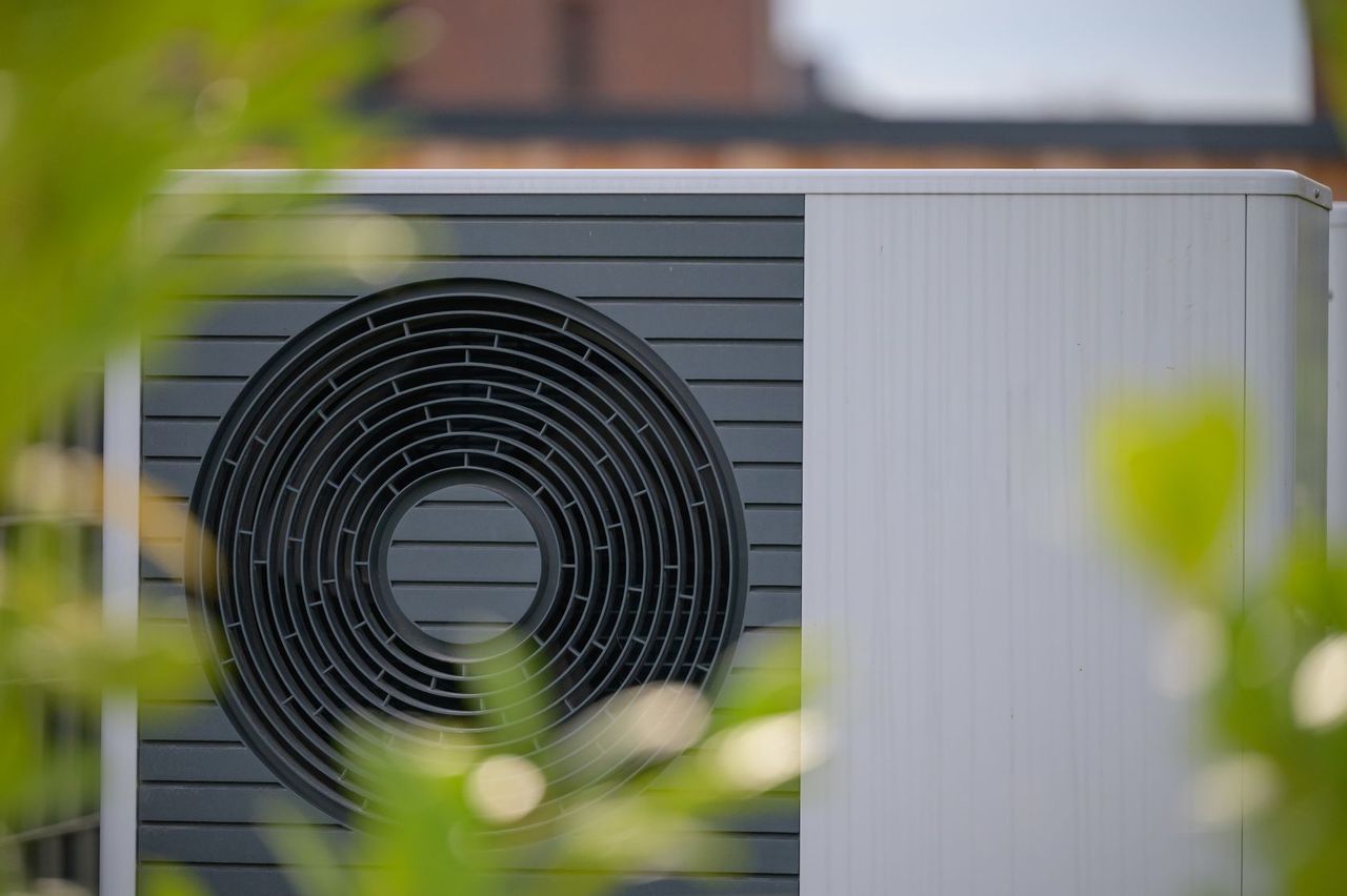 Nahaufnahme einer runden Lüftungsöffnung einer Wärmepumpe an einem grau-weißen Bauwerk, die im Vordergrund teilweise von verschwommenen grünen Blättern verdeckt wird.