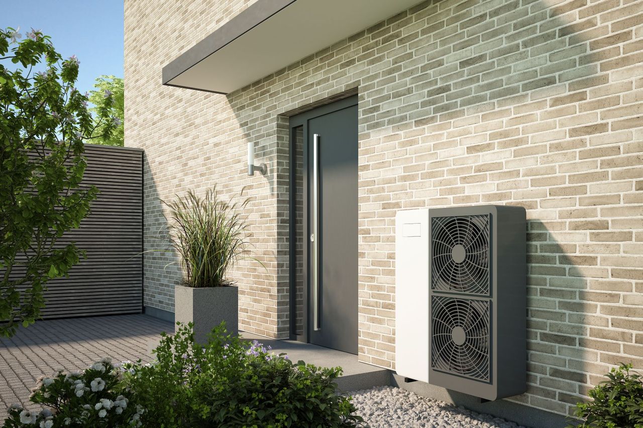 Fassade eines modernen Hauses mit Backsteinmauern, einer grauen Tür, einer Wärmepumpeneinheit, einer Topfpflanze und Grünpflanzen im Sonnenlicht.