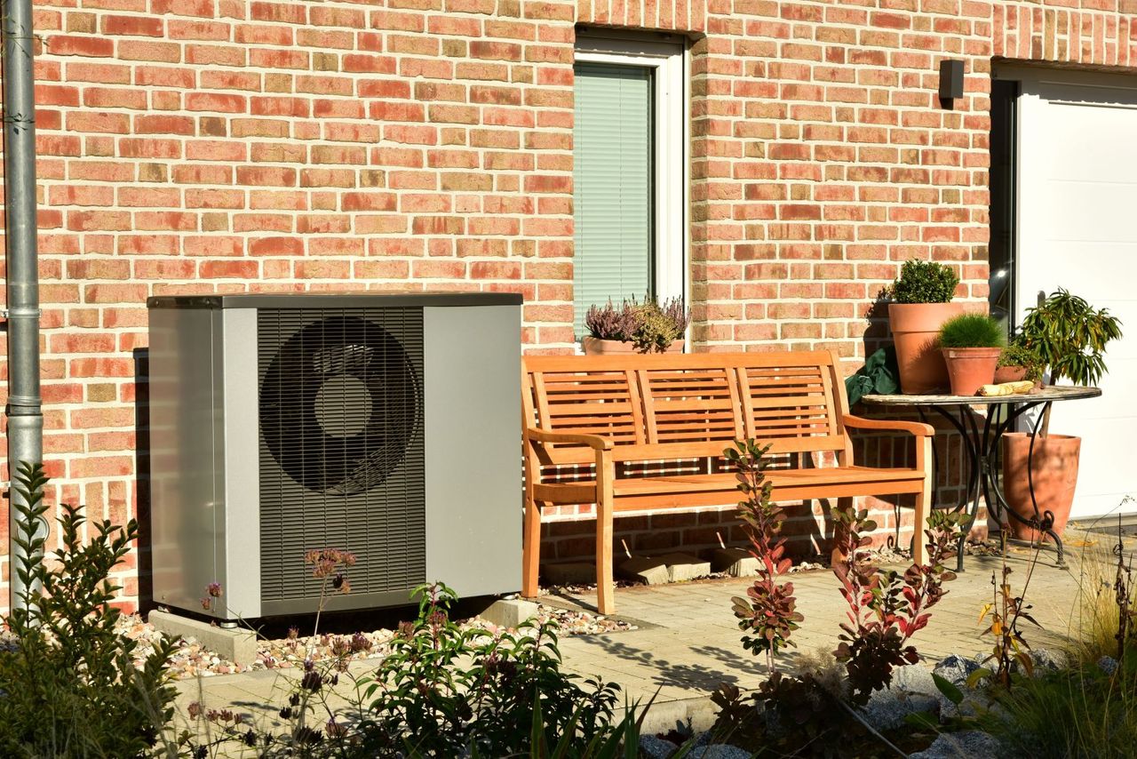 Eine Terrasse aus Ziegelsteinen mit einer Holzbank, Topfpflanzen und einer Außenklimaanlage neben einem Haus.