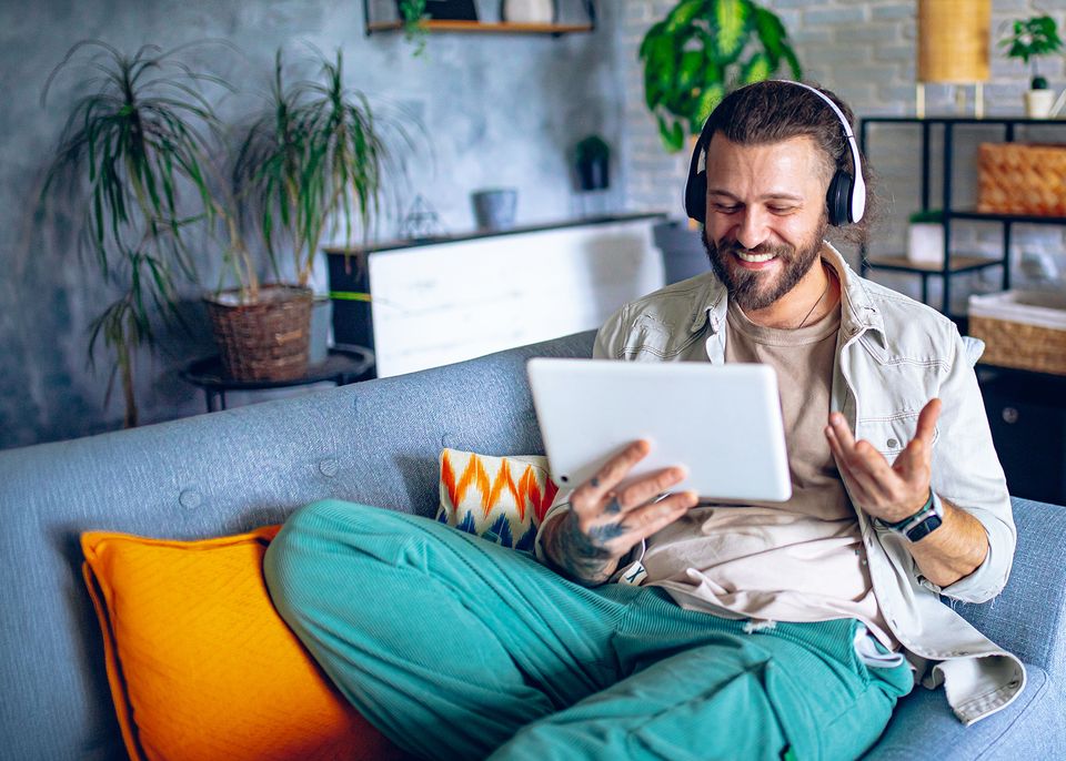 Glasfaser: Eine Mann mit Kopfhörern sitzt auf einem grauen Sofa, umgeben von bunten Kissen und Pflanzen, und lächelt, während er über ein Tablet per Videochat spricht.