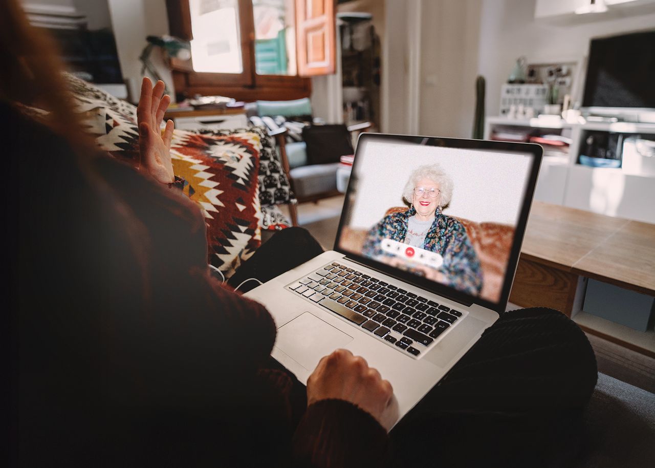 Frau sitzt mit Laptop auf dem Sofa. Dieser zeigt einen Videocall mit der Großmutter.