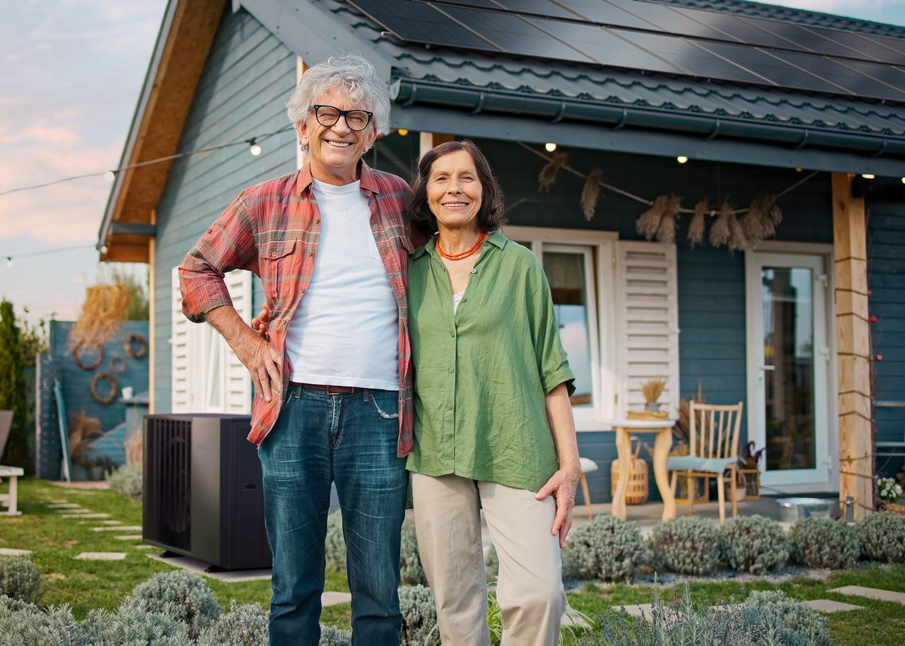 Älteres Ehepaar steht glücklich vor einem Haus mit PV-Anlage und Wärmepumpe (KI generiert)
