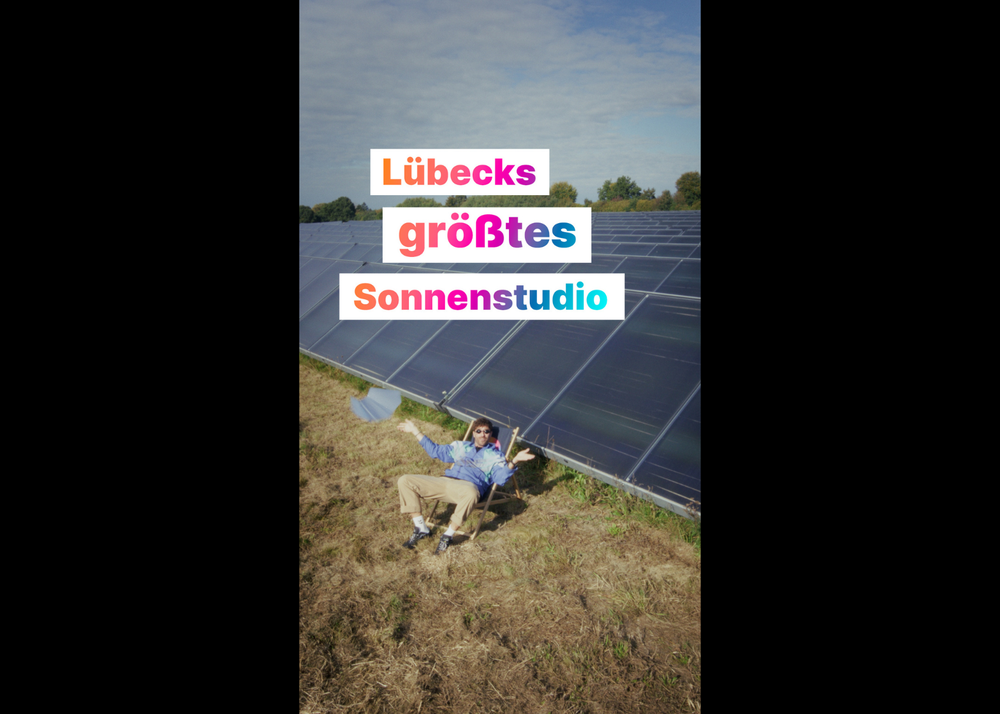 Eine Person, die auf einem Stuhl neben Sonnenkollektoren auf einem Feld unter einem Schild mit der Aufschrift „Lübecks größtes Sonnenstudio“ in bunten Buchstaben faulenzt.