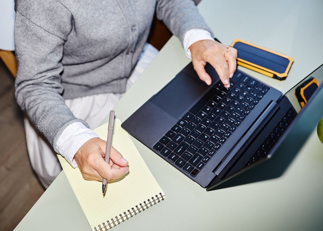 Person in einem grauen Pullover, die einen Laptop benutzt und einen Stift über einen Notizblock auf einem Schreibtisch hält, mit einem Smartphone in der Nähe.