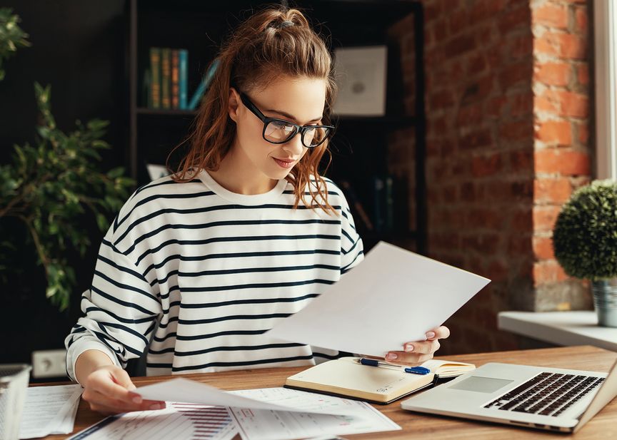 Eine Frau in einem gestreiften Pullover arbeitet an einem Schreibtisch mit Papieren und einem Laptop, umgeben von Büchern und Pflanzen in einem gemütlichen Raum.