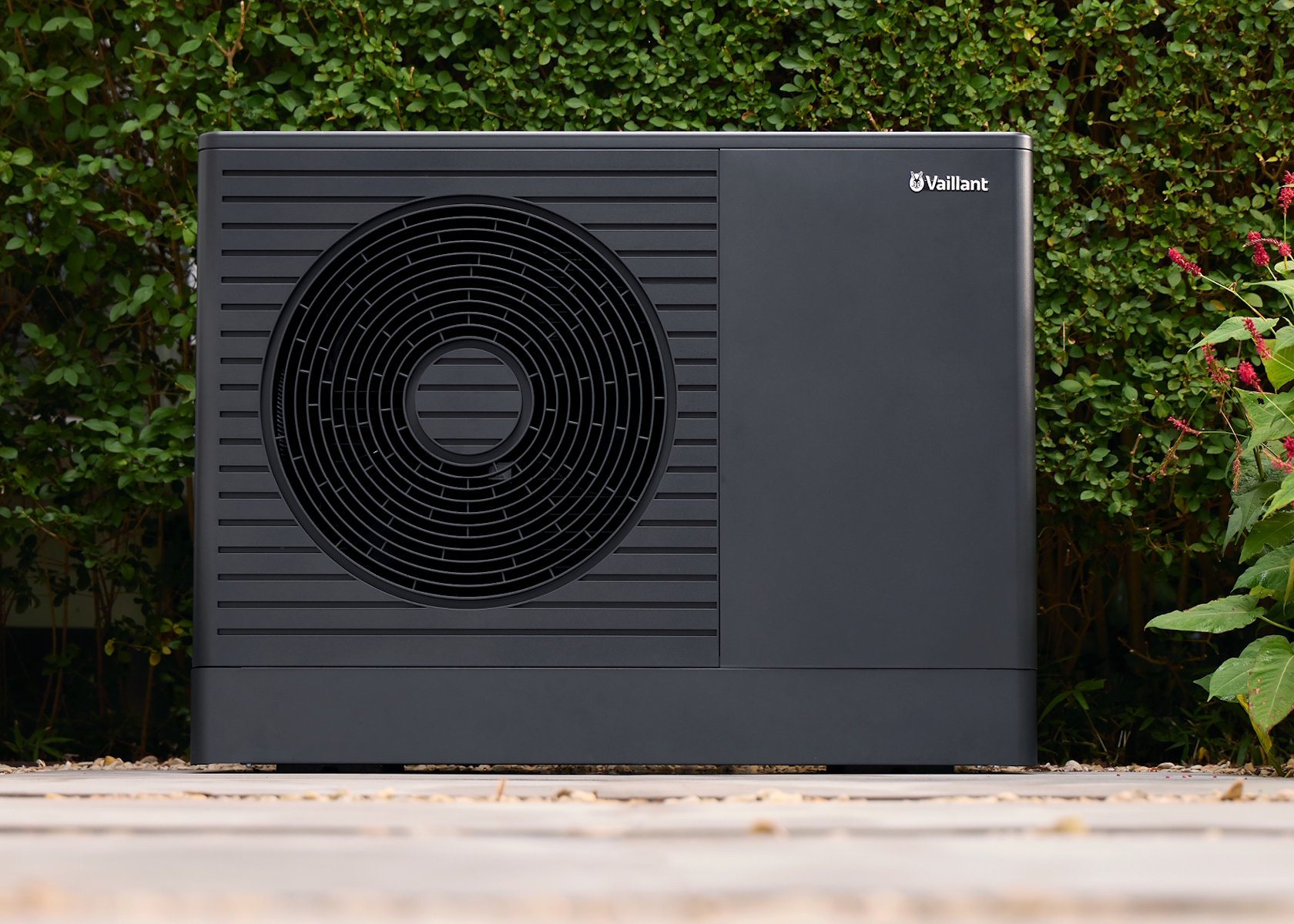 Eine schwarze Wärmepumpeneinheit für den Außenbereich mit rundem Lüfterdesign vor dem Hintergrund von grünem Laub und rosa Blüten. (Lizenz: Vaillant)