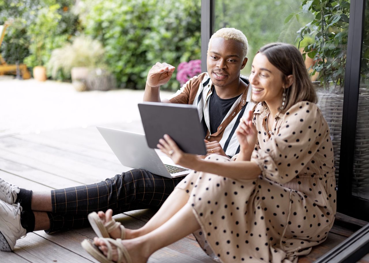 Zwei Personen sitzen auf einer Holzterrasse, unterhalten sich und schauen dabei auf ein Tablet und einen Laptop. Im Hintergrund ist üppiges Grün zu sehen.