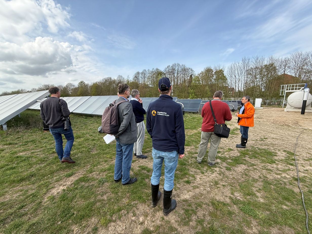 Eine Gruppe von Menschen in Outdoor-Kleidung unterhält sich in der Nähe von Sonnenkollektoren auf einer Wiese unter teilweise bewölktem Himmel.