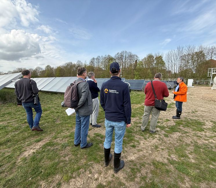 Eine Gruppe von Menschen in Outdoor-Kleidung unterhält sich in der Nähe von Sonnenkollektoren auf einer Wiese unter teilweise bewölktem Himmel.