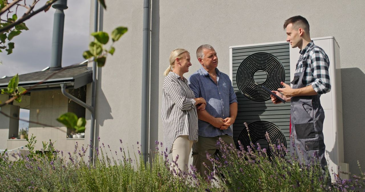 Ein Techniker erklärt einem Paar vor ihrem Haus, umgeben von Lavendelpflanzen, wie man eine Wärmepumpe bedient.