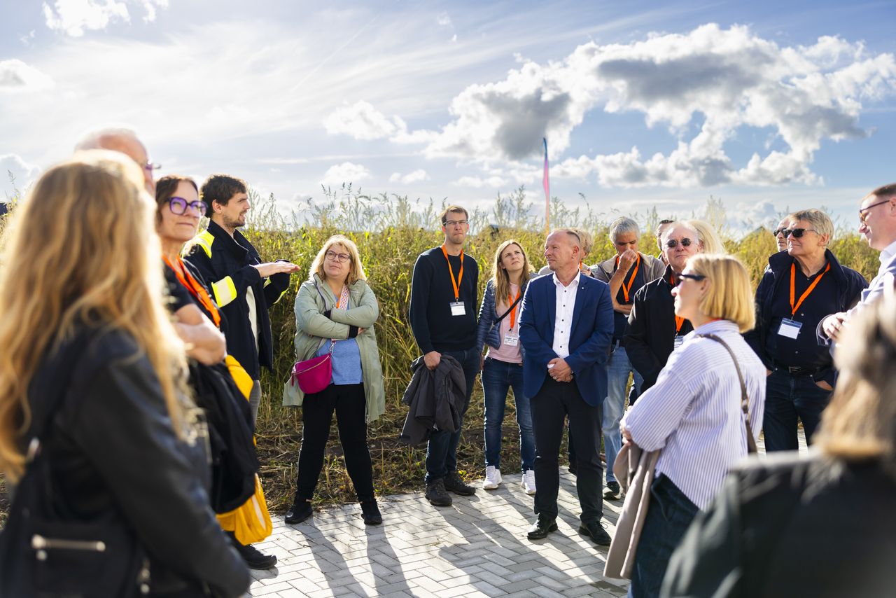 An einem sonnigen Tag versammelte sich eine Gruppe von Menschen im Freien; sie hörten aufmerksam zu, trugen Umhängebänder und Namensschilder, vor dem Hintergrund von Schilf und Wolken.
