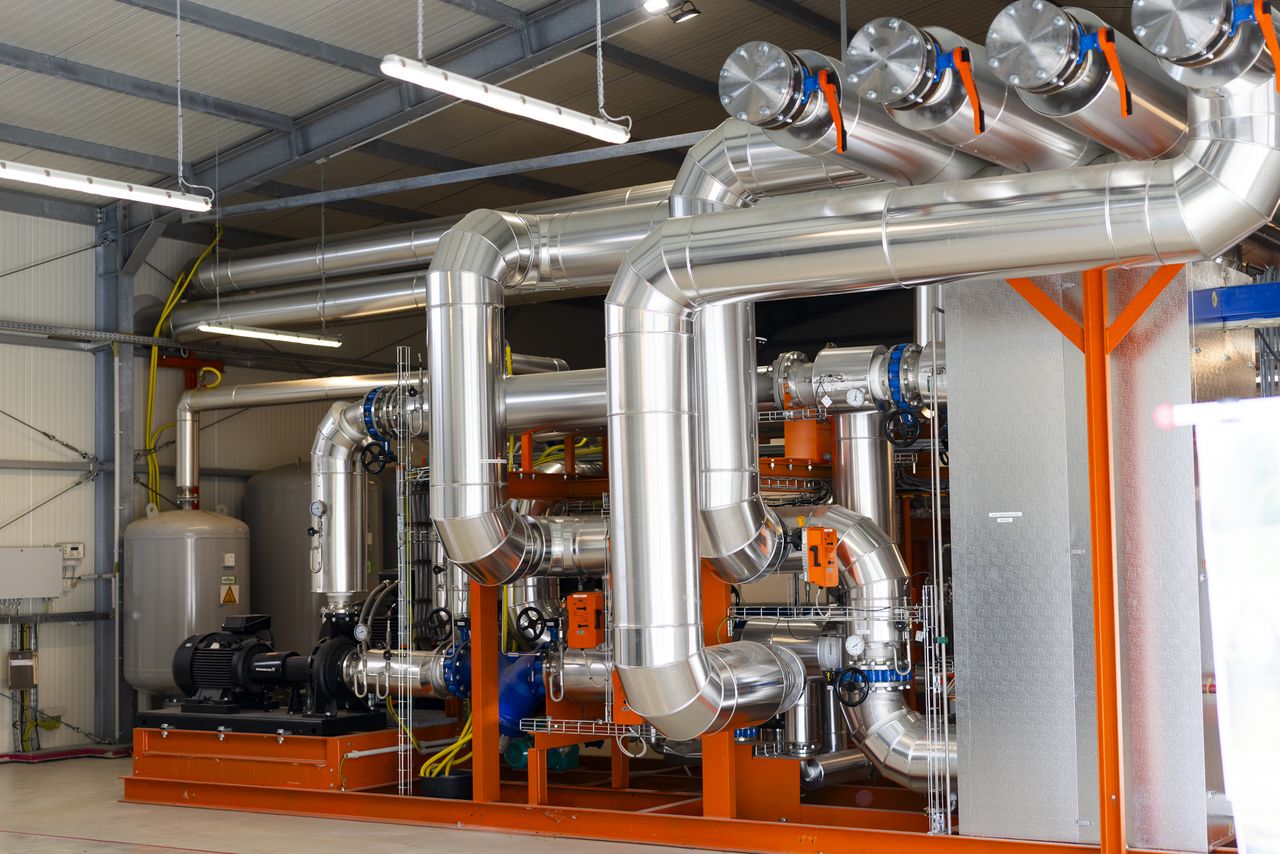 Industrielles Rohrleitungssystem mit großen Edelstahlrohren, orangefarbenen Halterungen und Messgeräten im Inneren einer Anlage.