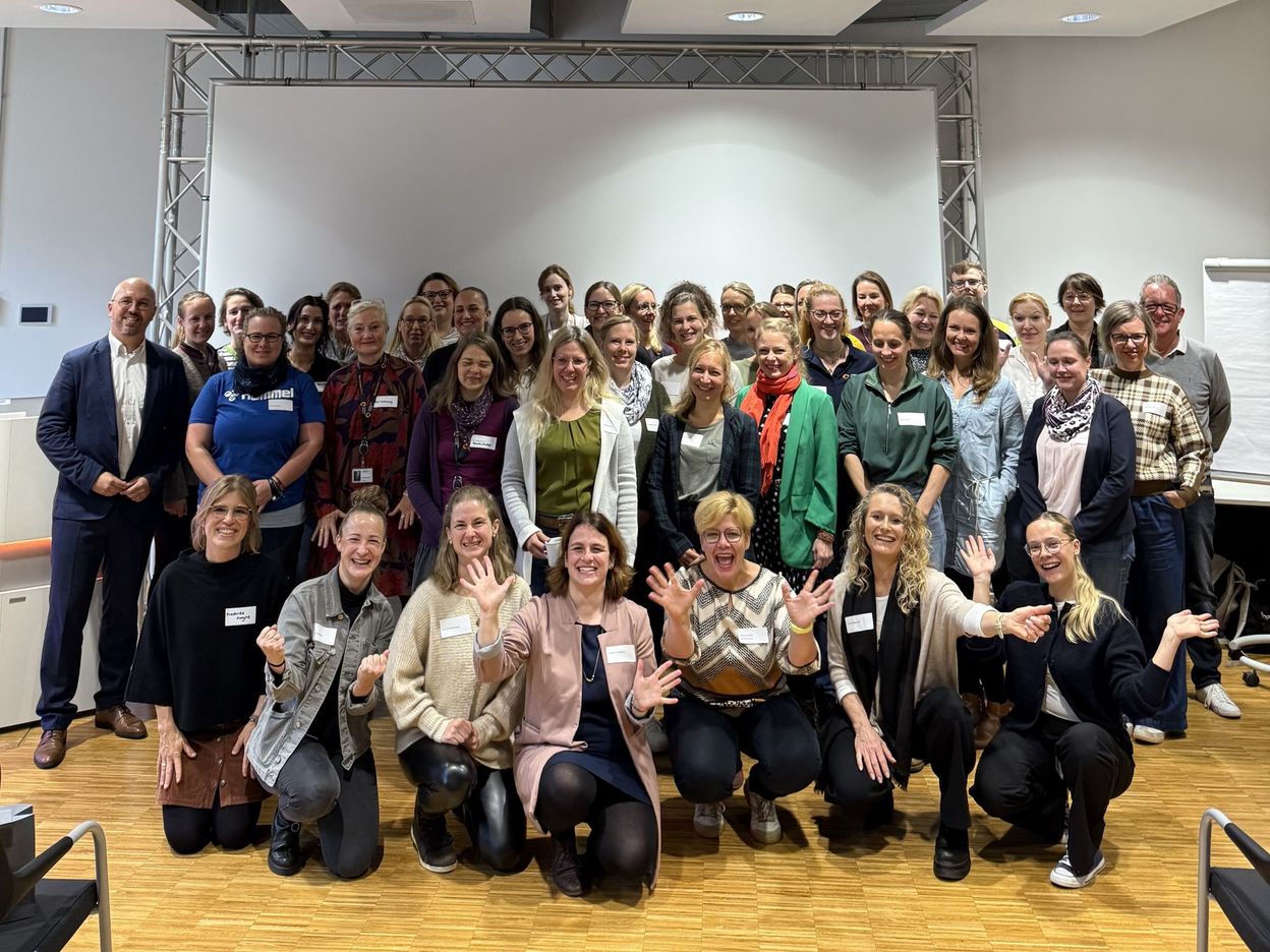 Eine Gruppe von Menschen, überwiegend Frauen, die in einem Raum vor einer weißen Leinwand lächeln und gemeinsam posieren. Einige knien in der ersten Reihe.