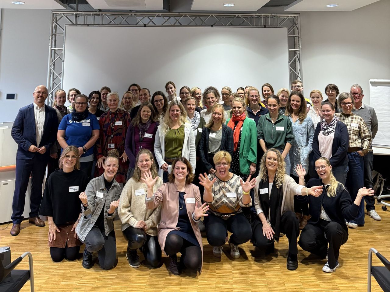 Eine Gruppe von Menschen, überwiegend Frauen, die in einem Raum vor einer weißen Leinwand lächeln und gemeinsam posieren. Einige knien in der ersten Reihe.