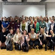Eine Gruppe von Menschen, überwiegend Frauen, die in einem Raum vor einer weißen Leinwand lächeln und gemeinsam posieren. Einige knien in der ersten Reihe.