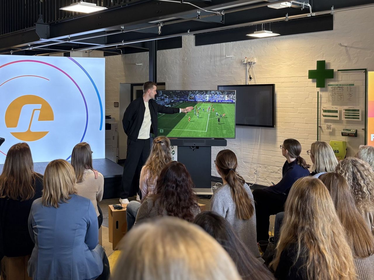 Ein Referent hält vor einer Gruppe von Frauen, die in einem Raum sitzen, einen Vortrag, während auf einer Leinwand eine Fußballspiel zu sehen ist.