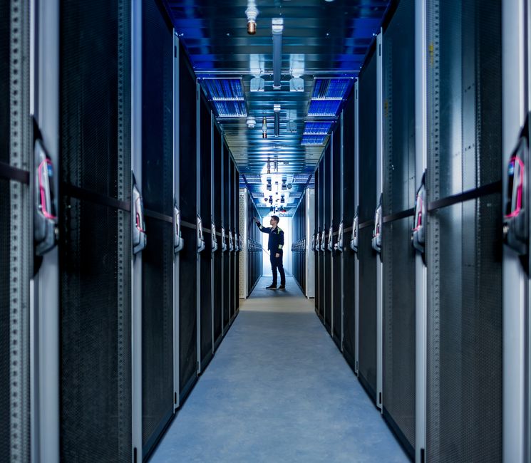 Eine Person arbeitet in einem schmalen Gang zwischen Reihen von Server-Racks in einem hell erleuchteten Rechenzentrum.