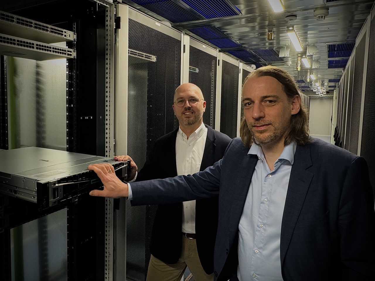 Jens Meier und Christoph Schweizer in Anzügen stehen in einem Serverraum; einer von ihnen berührt ein Serverrack, im Hintergrund sind Reihen von Geräten zu sehen.