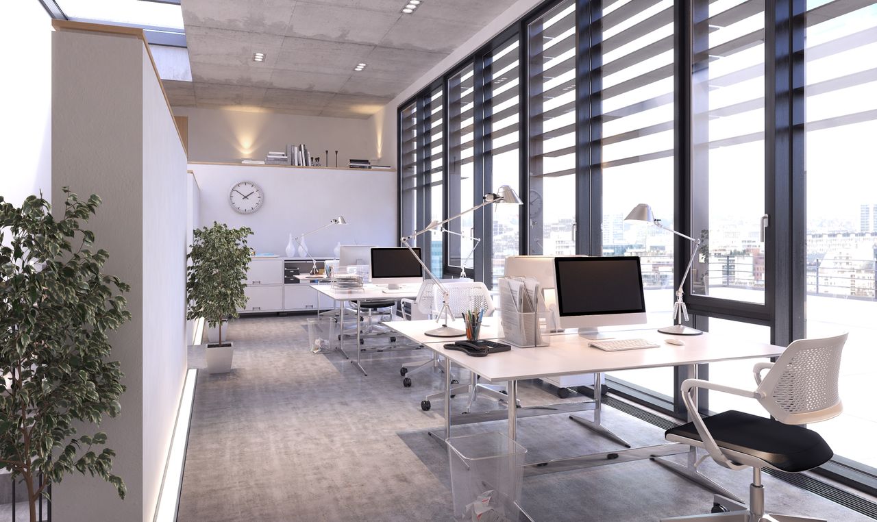 Modern office space with large windows, sleek desks, computers, chairs, and potted plants. Natural light brightens the room, creating an airy feel.