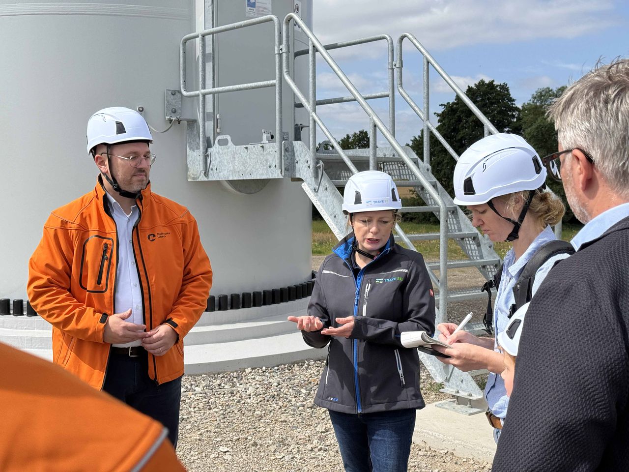Eine Gruppe von Personen, die Schutzhelme und Schutzjacken tragen, steht in der Nähe eines Industriegebäudes und unterhält sich, wobei eine Person Notizen macht.