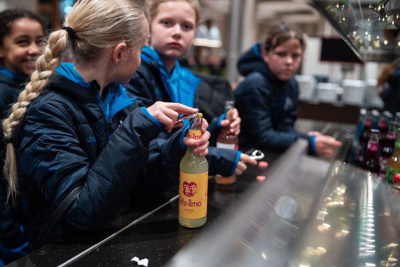 Children in blue jackets gather around a counter, opening bottles of drinks, with various beverages displayed in the foreground.