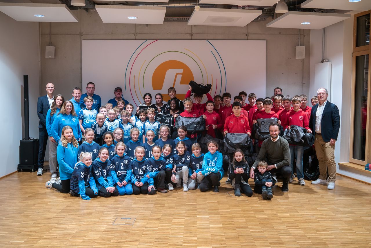 A large group of children and adults in sports uniforms pose together in a room with a logo on the wall. Some hold bags, and everyone is smiling.