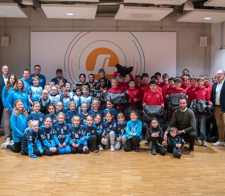 A large group of children and adults in sports uniforms pose together in a room with a logo on the wall. Some hold bags, and everyone is smiling.