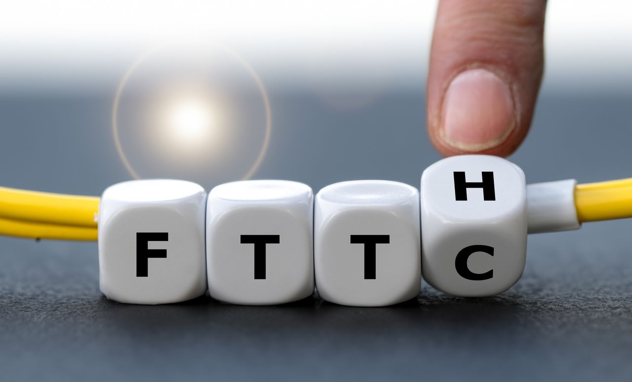 Vor einem unscharfen Hintergrund dreht eine Hand einen weißen Würfel von „FTTC“ auf „FTTH“, unter dem Würfel ist ein gelbes Kabel zu sehen (KI generiert)