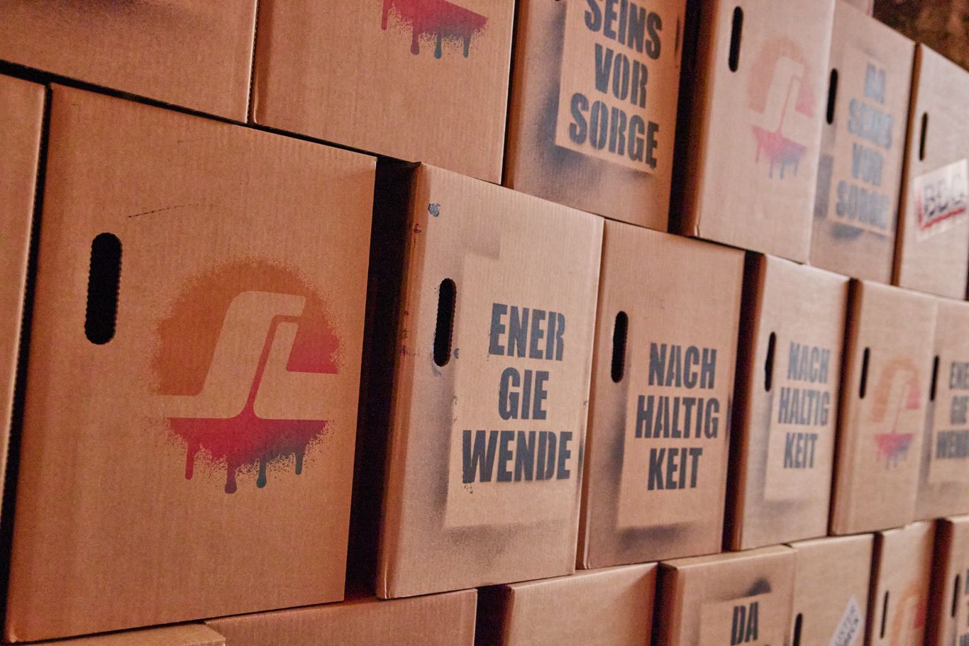 Stacked cardboard boxes with German text and a logo on one, featuring phrases like "ENERGIE WENDE" and "NACH HALTIG KEIT."