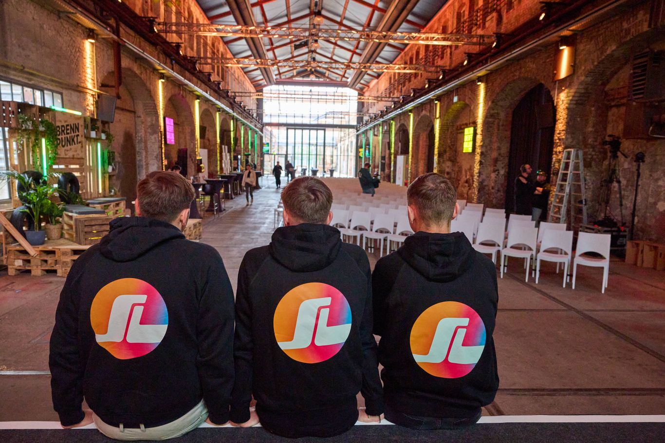 Three people wearing matching hoodies with colorful logos sit facing an empty event space with rows of white chairs and industrial decor.