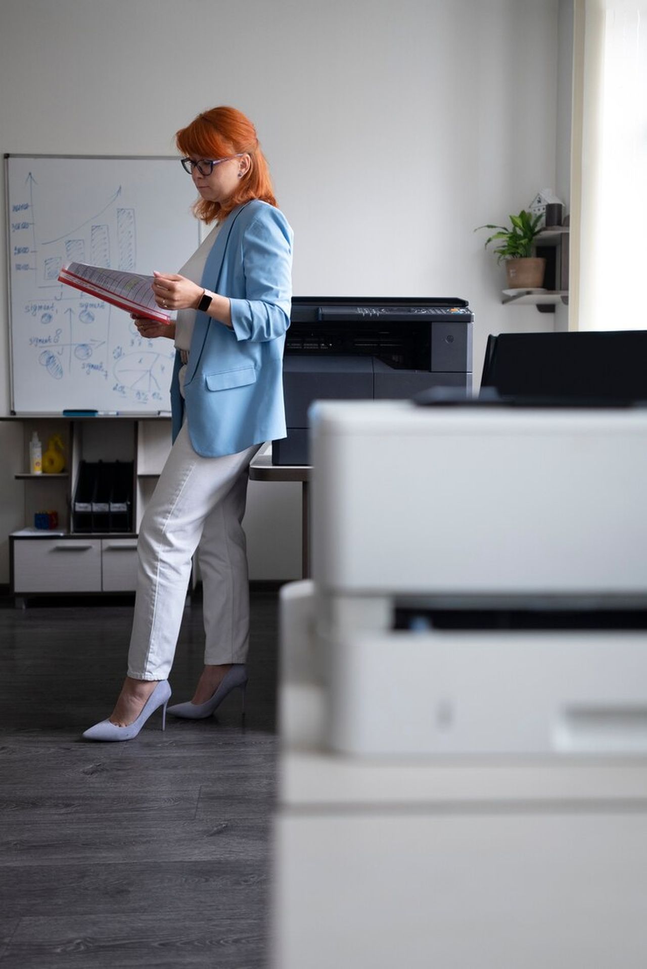 A woman with red hair, wearing a blue blazer and white pants, holds a folder while standing in an office with a whiteboard and printer.