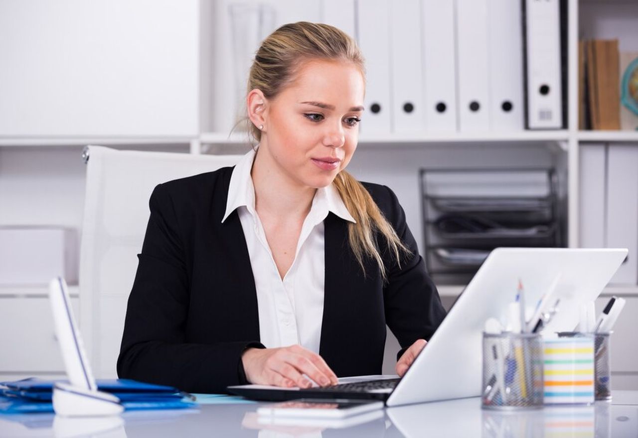 Woman in a black blazer working on a laptop at a desk, surrounded by office supplies and shelves in the background.